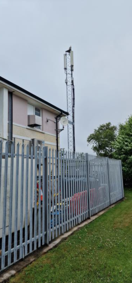 A photograph showing an existing telecommunications lattice tower standing behind a grey metal security fence next to a building.
