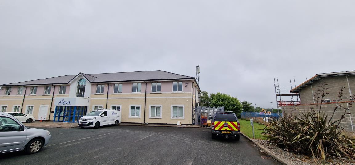 Street-level photograph showing a two-story commercial building with 'Argon' signage, a parking area with vehicles, and a telecommunications mast visible in the background.