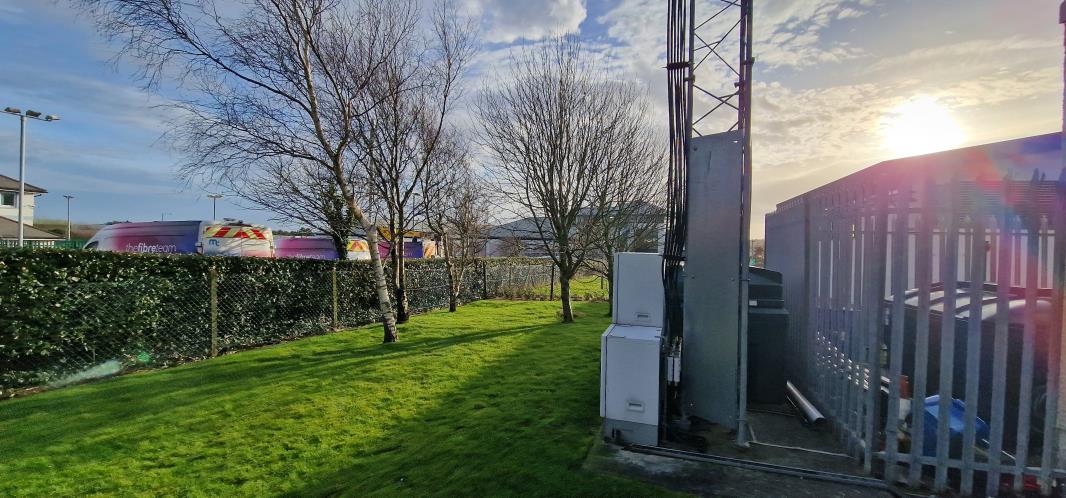 A photograph showing the existing telecommunications site with a lattice tower, white equipment cabinets, and fencing adjacent to a grassy area and road.