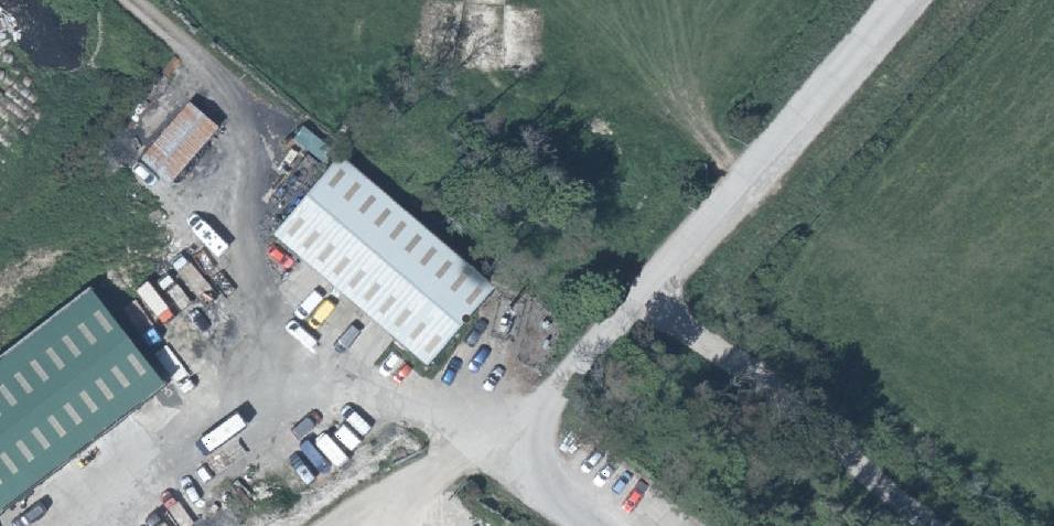 An aerial view of a commercial or industrial site featuring large warehouse buildings, a vehicle yard with numerous parked vans, and surrounding green fields.