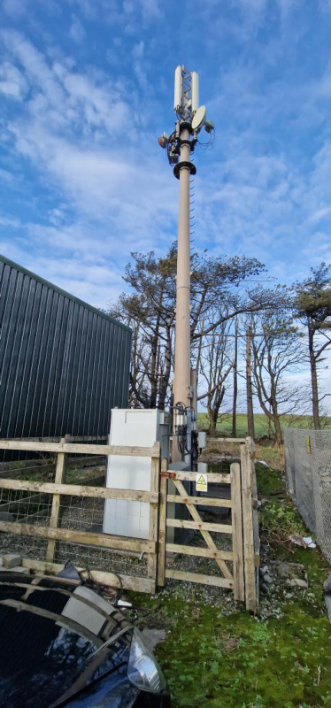 A photograph showing an existing telecommunications monopole mast with antennas, a white equipment container, and a wooden boundary fence in a rural setting.
