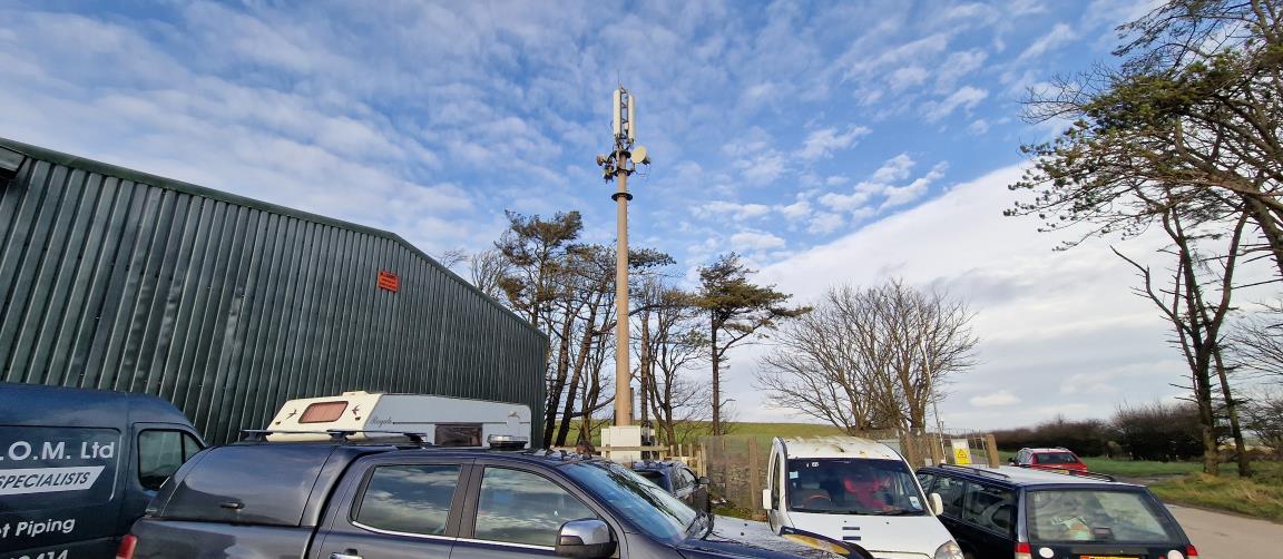 A photograph showing an existing telecommunications monopole mast with antennas, situated next to a large green industrial building with vehicles parked in the foreground.