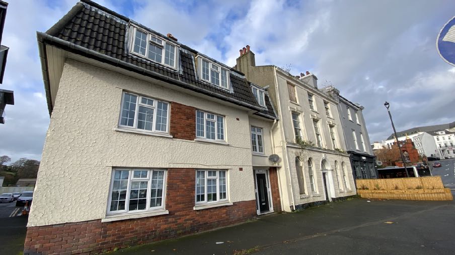 A street-level photograph showing the existing buildings on the site, featuring a white rendered building with dormer windows adjacent to older terraced structures with arched ground-floor windows.