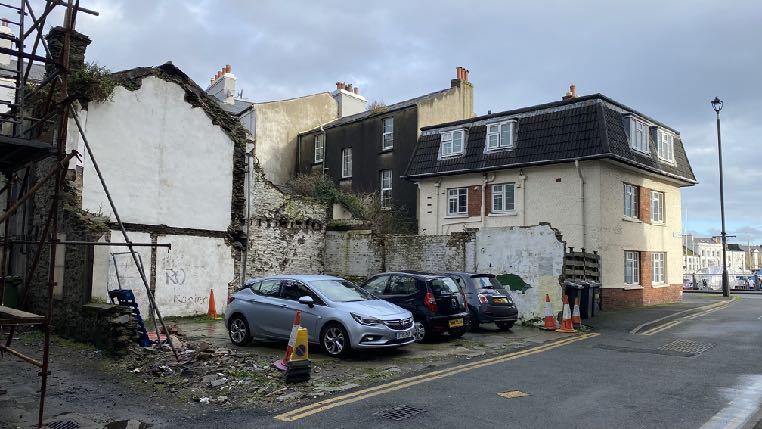 A street-level photograph showing a partially demolished building with scaffolding and debris on the left, adjacent to a fully intact two-story residential house on the right.