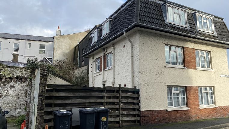 A photograph showing the exterior of a two-story residential house with white rendering and brick accents, situated behind a wooden fence and waste bins.