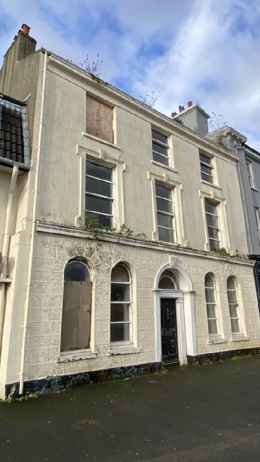 A photograph of a derelict, two-story white building with boarded windows and vegetation growing from the roofline, likely a former commercial or residential property.