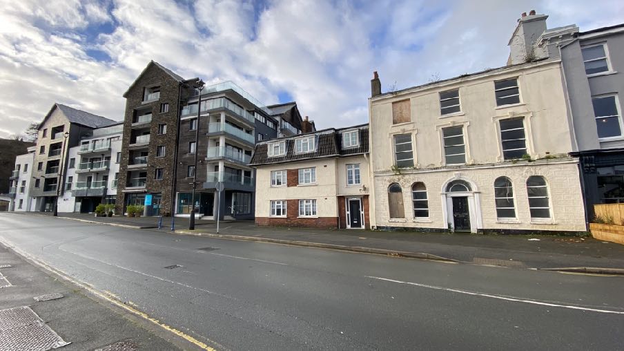 A street-level photograph showing a row of buildings in Douglas, featuring modern apartment blocks on the left and an older white terraced building with boarded windows on the right.