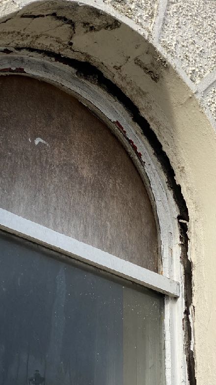 A close-up photograph showing the deteriorating condition of an arched window or doorway, featuring significant cracking in the render and peeling paint.