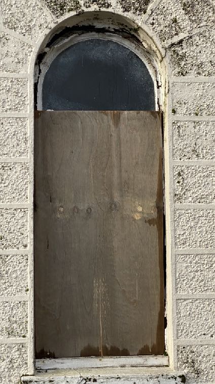 A close-up photograph of a boarded-up arched window or door opening on a roughcast stone wall, likely part of a building scheduled for demolition.