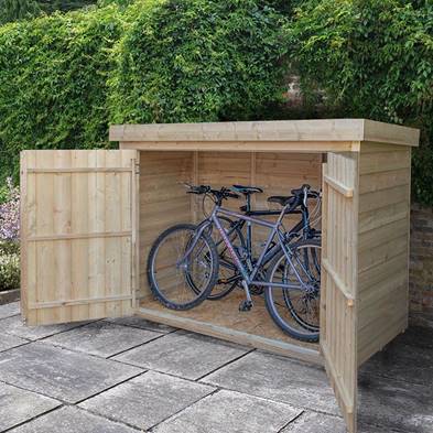 A wooden cycle storage shed with double doors open, revealing several bicycles parked inside on a paved surface with a hedge in the background.