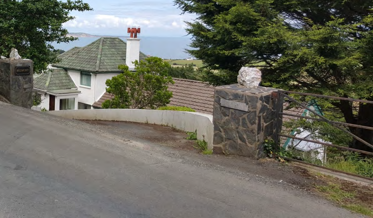 A street-level photograph showing a white detached house with a green roof, stone gateposts, and a view of the sea in the distance.