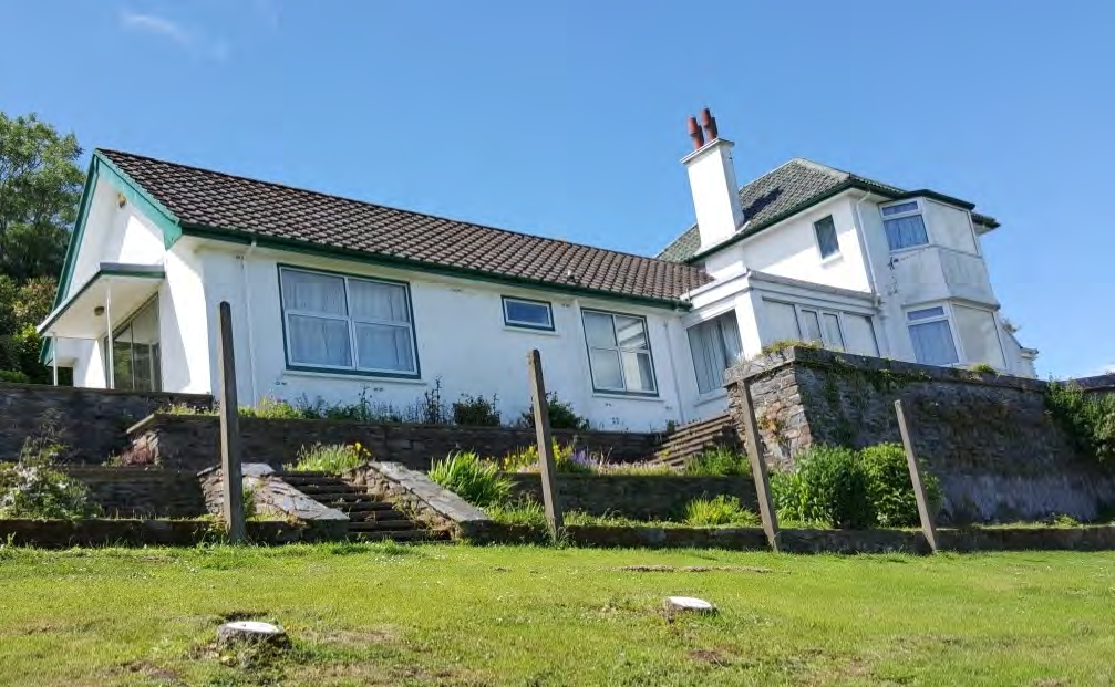 A white detached house with a tiled roof and green trim is situated on a sloping site with stone retaining walls and steps.