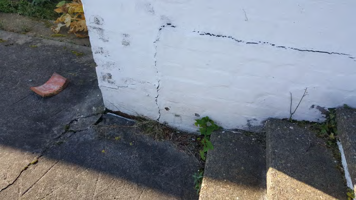A close-up photograph showing significant vertical and horizontal cracking in a white painted brick wall and adjacent concrete paving with weeds.