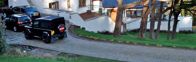 A high-angle photograph showing a paved driveway with several parked vehicles, including a Land Rover, leading up to a modern white detached house with wooden accents and surrounding landscaping.