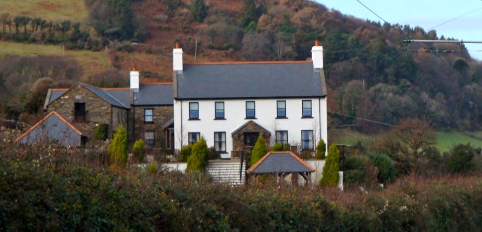 A photograph showing a large white detached house with a slate roof situated on a hillside, featuring white chimneys and stone outbuildings to the left.