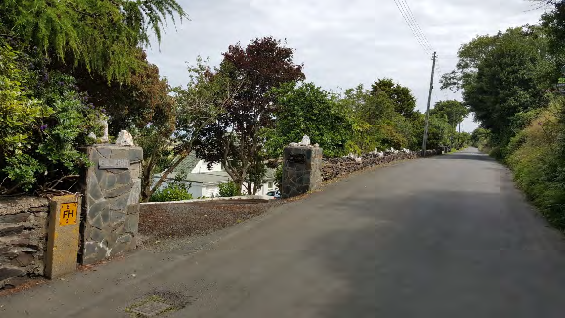 A photograph showing a rural road entrance marked by stone pillars and a gravel driveway leading to a white house.