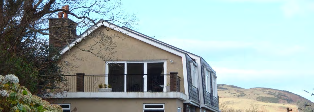 A photograph of a modern, two-story detached house featuring a balcony and large windows, situated in a rural environment with a hill in the background.