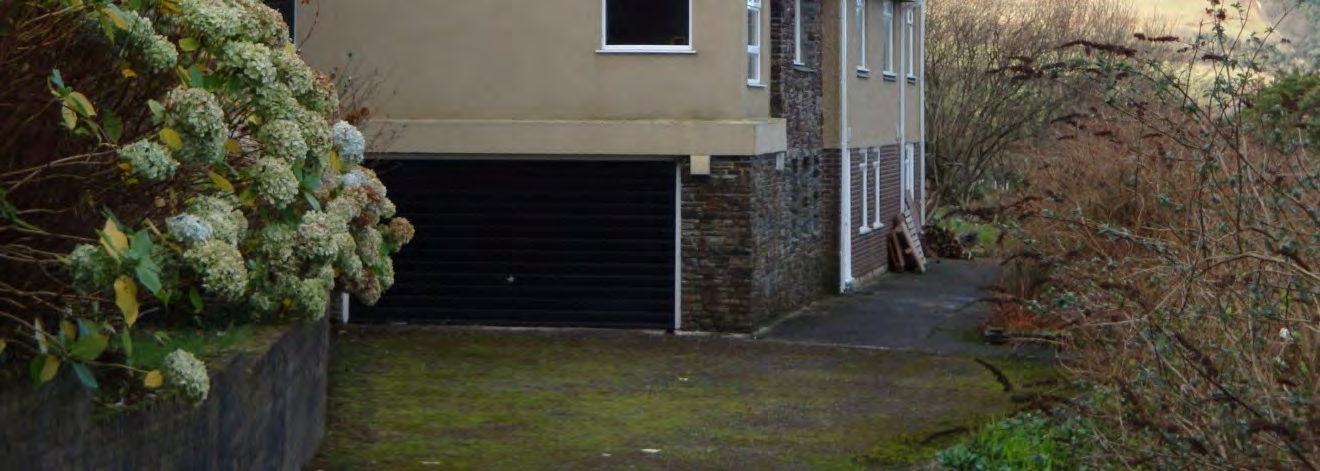 A photograph showing the side elevation of a residential property featuring a black roller shutter garage door, stone and rendered walls, and a paved driveway area surrounded by vegetation.