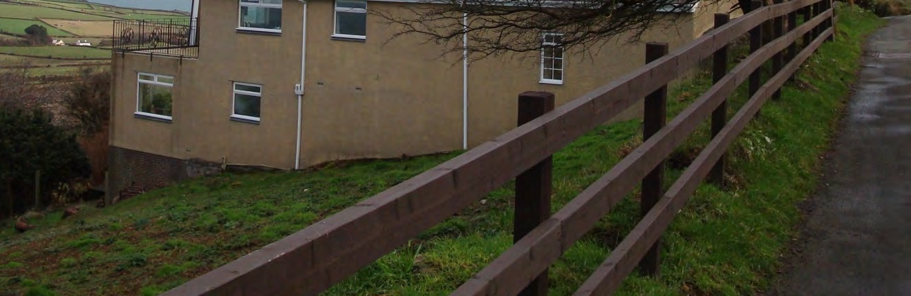 A photograph showing the exterior of a two-story building with a tan rendered finish and white windows. A wooden fence runs along the foreground next to a paved path, with rural fields visible in the background.