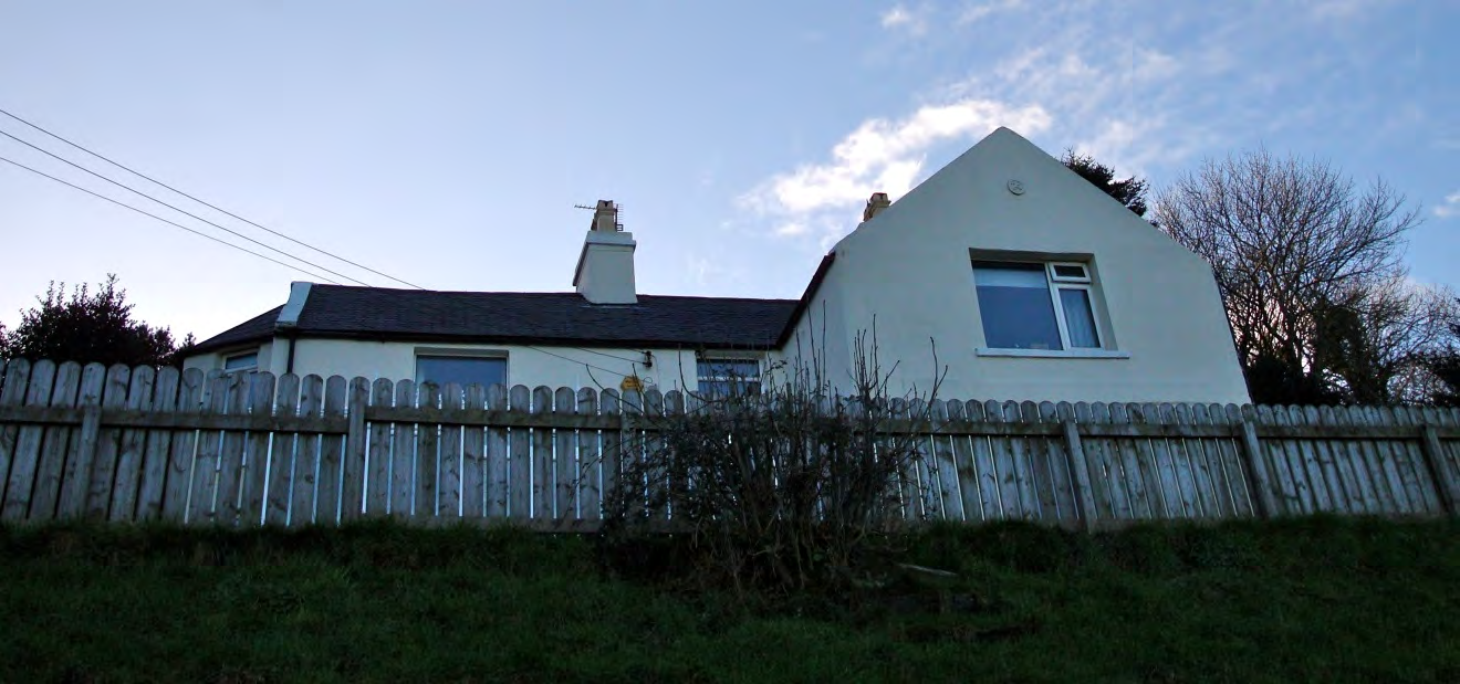 A low-angle photograph of a white, single-story detached house with a dark roof, situated behind a wooden fence in a grassy area.