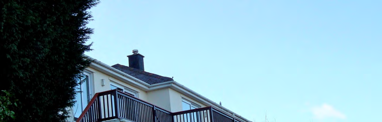 A photograph showing the upper portion of a detached house with a tiled roof, chimney, and a wooden deck railing against a clear blue sky.