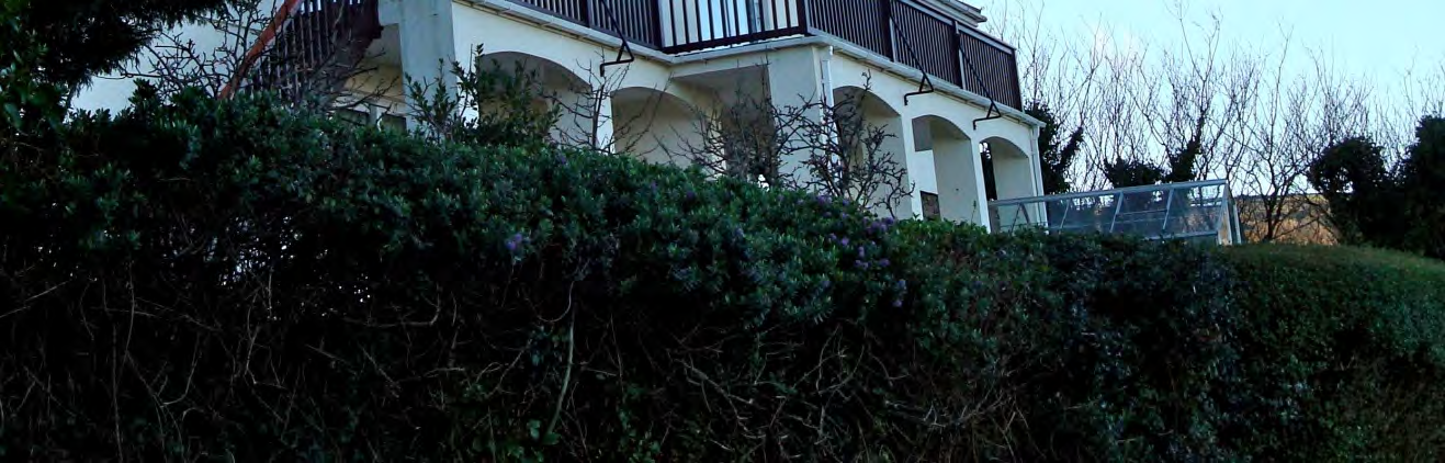 A photograph showing a white residential building with arched ground floor openings and an upper balcony, partially obscured by a large hedge in the foreground.