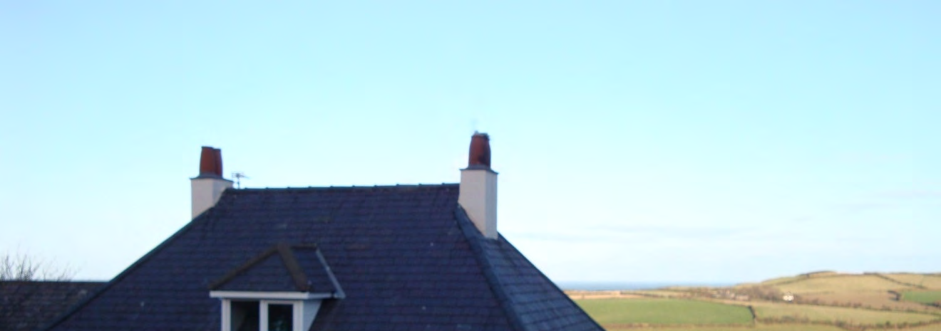 A photograph showing the slate roof of a house featuring two chimneys and a dormer window. The background displays a rural landscape with green fields and a distant horizon.