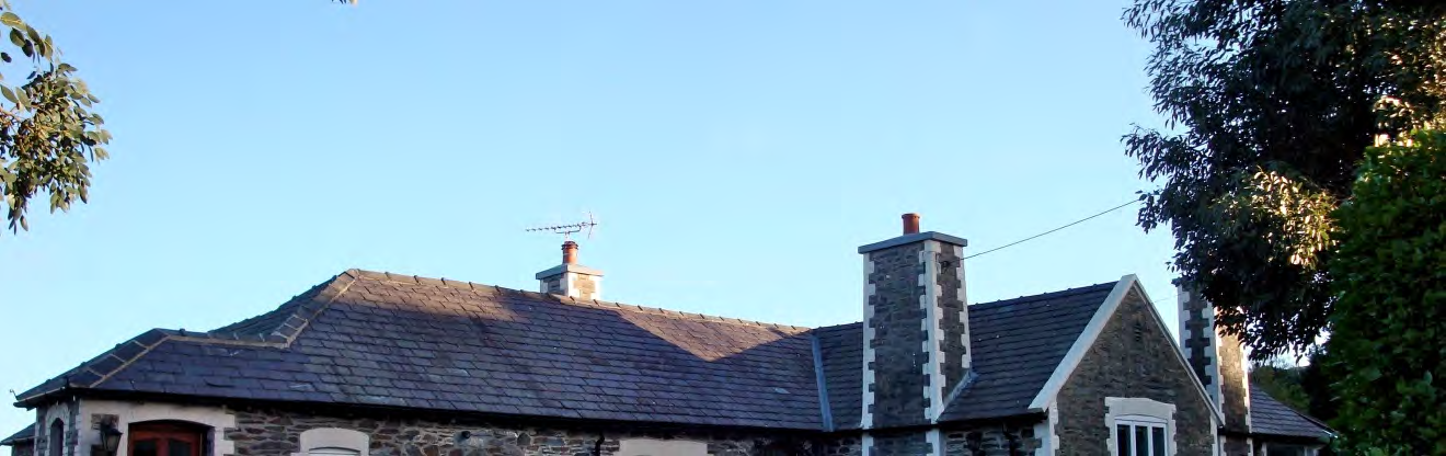 A photograph showing the roofline and upper facade of a traditional stone building with slate roofing and chimneys against a blue sky.