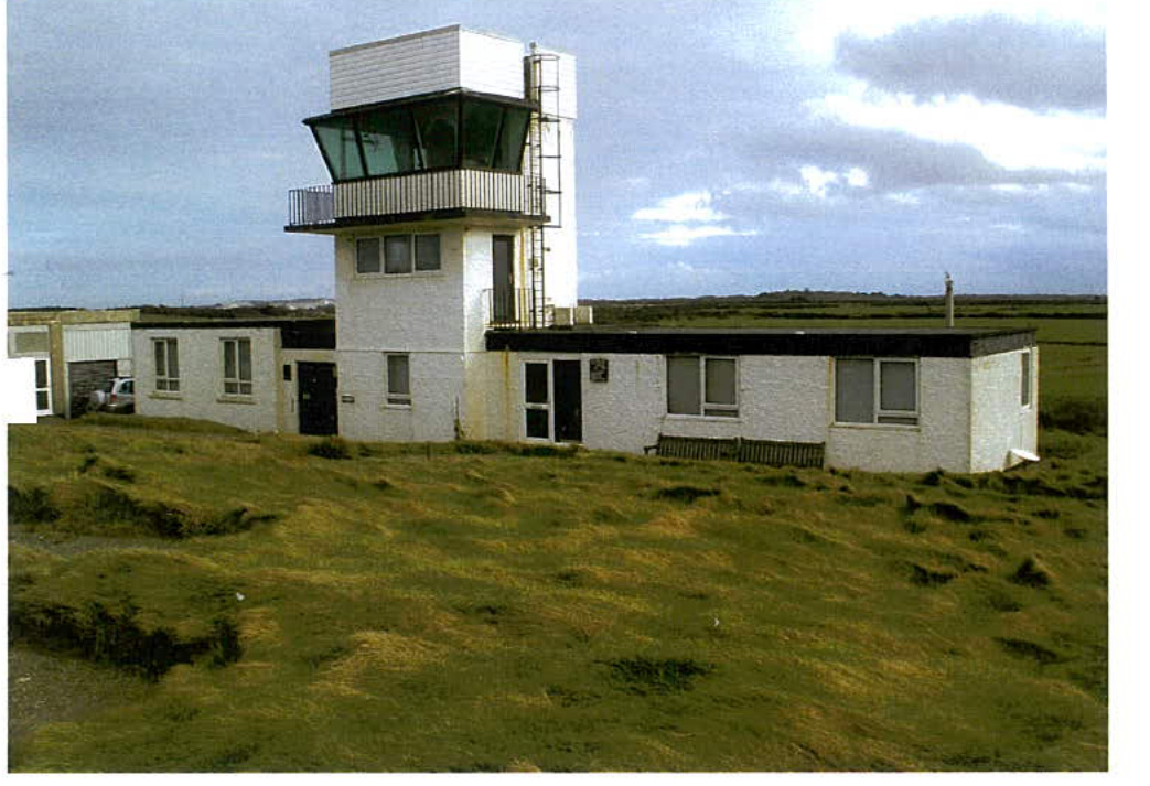 A photograph showing a white building complex with a prominent tower featuring a glass observation deck, situated in a grassy rural field.