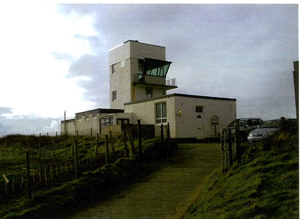 A photograph showing a white, modern building with a prominent tower section and a lower single-story extension, situated on a grassy slope with a driveway.