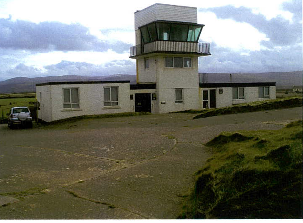 A photograph showing a white building with a prominent control tower structure, situated in a rural setting with hills in the distance.