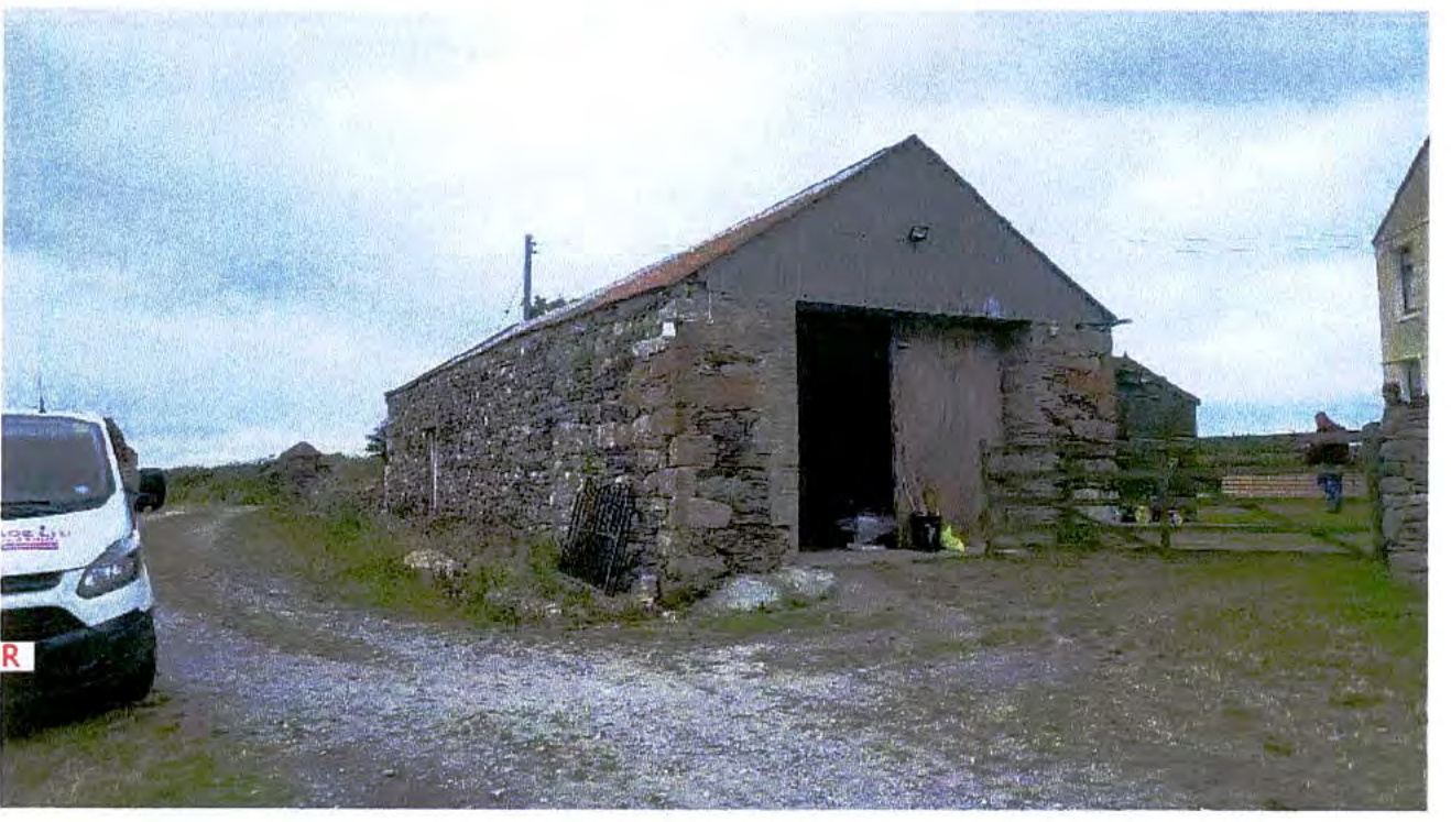 A photograph showing a stone barn or outbuilding with a large open doorway, situated on a gravel driveway in a rural setting.