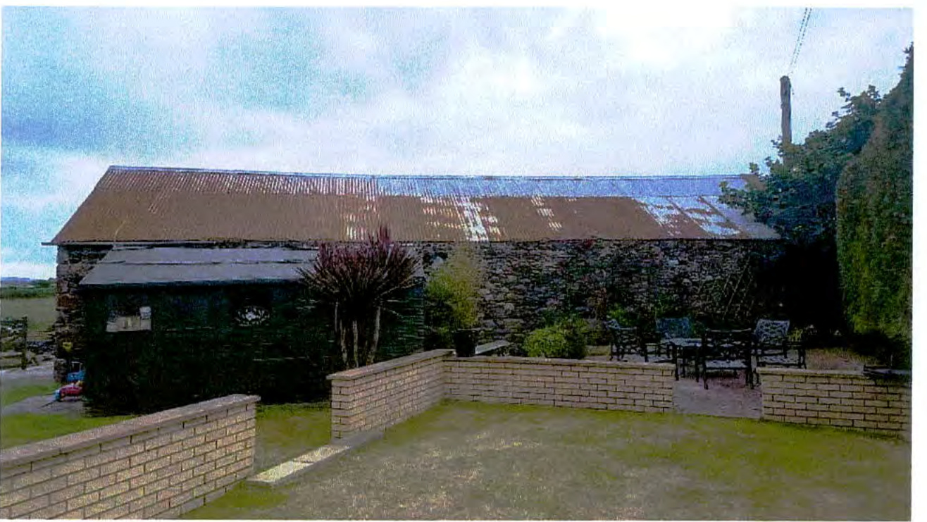A photograph showing a stone barn-like building with a corrugated roof and a patio area with low brick walls in the foreground.