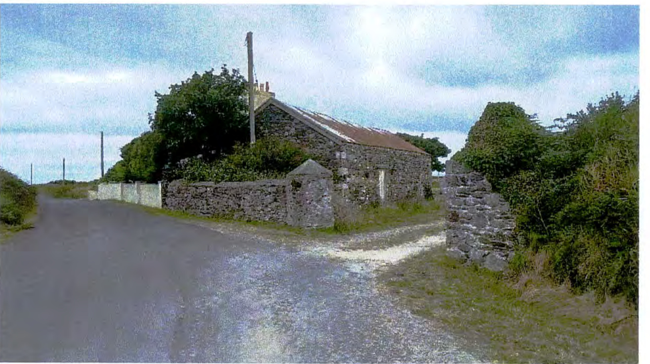 A photograph showing a stone building, likely a barn, situated beside a rural road with stone walls and vegetation.