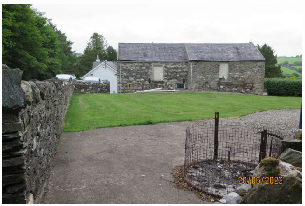 A photograph showing a large stone barn with boarded windows and a slate roof, situated next to a white building and stone walls.