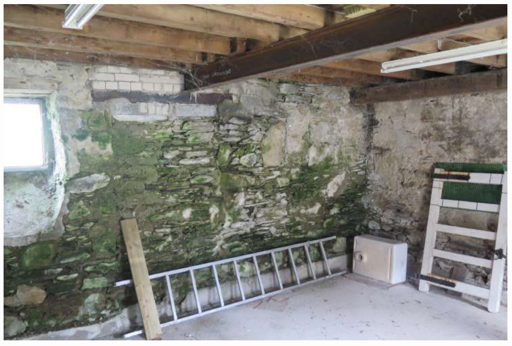 Interior photograph of a stone barn undergoing structural renovation, featuring exposed stone walls with moss, wooden joists, and a steel support beam.