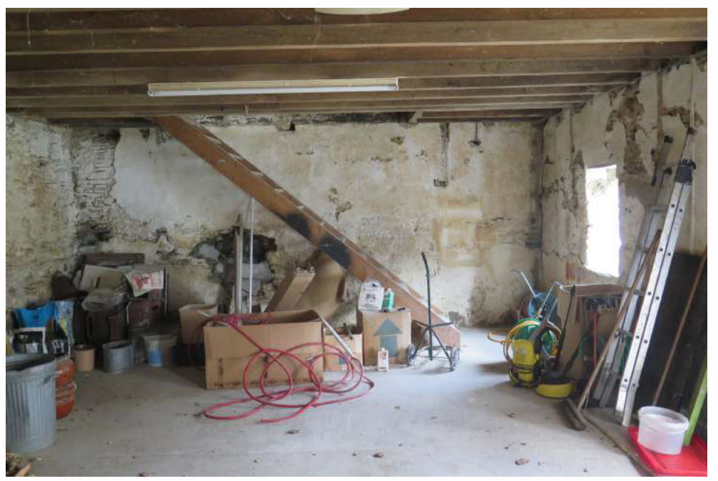 Interior photograph of a rustic barn or outbuilding showing exposed wooden ceiling joists and rough walls. The space is currently used for storage, containing items like a ladder, wheelbarrow, and boxes.