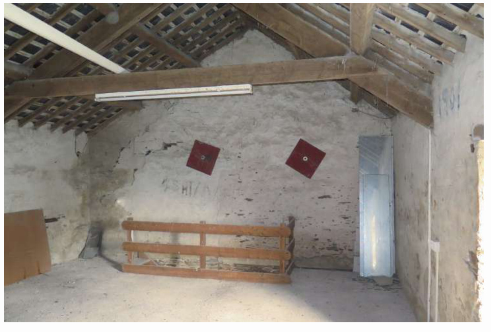 Interior photograph of a rustic barn space featuring exposed wooden roof beams, rough white walls, and a wooden barrier.