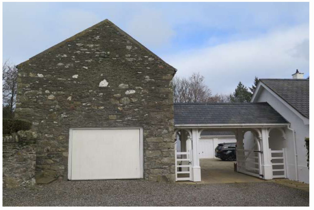 A photograph showing a stone barn building with a large white garage door next to a white wooden covered log store or carport.