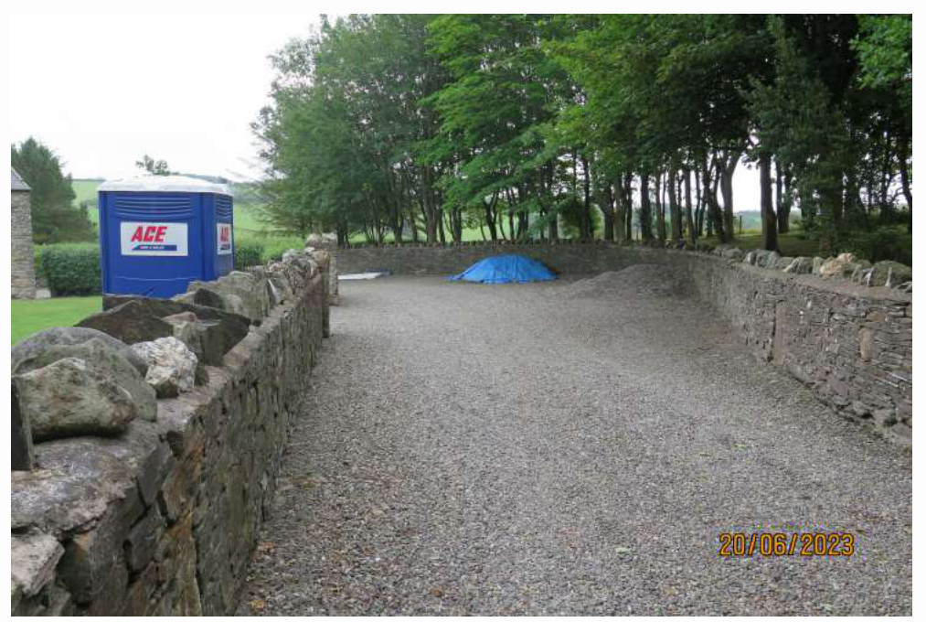 A photograph of a gravel yard or driveway enclosed by traditional dry stone walls, featuring a portable toilet on the left side.
