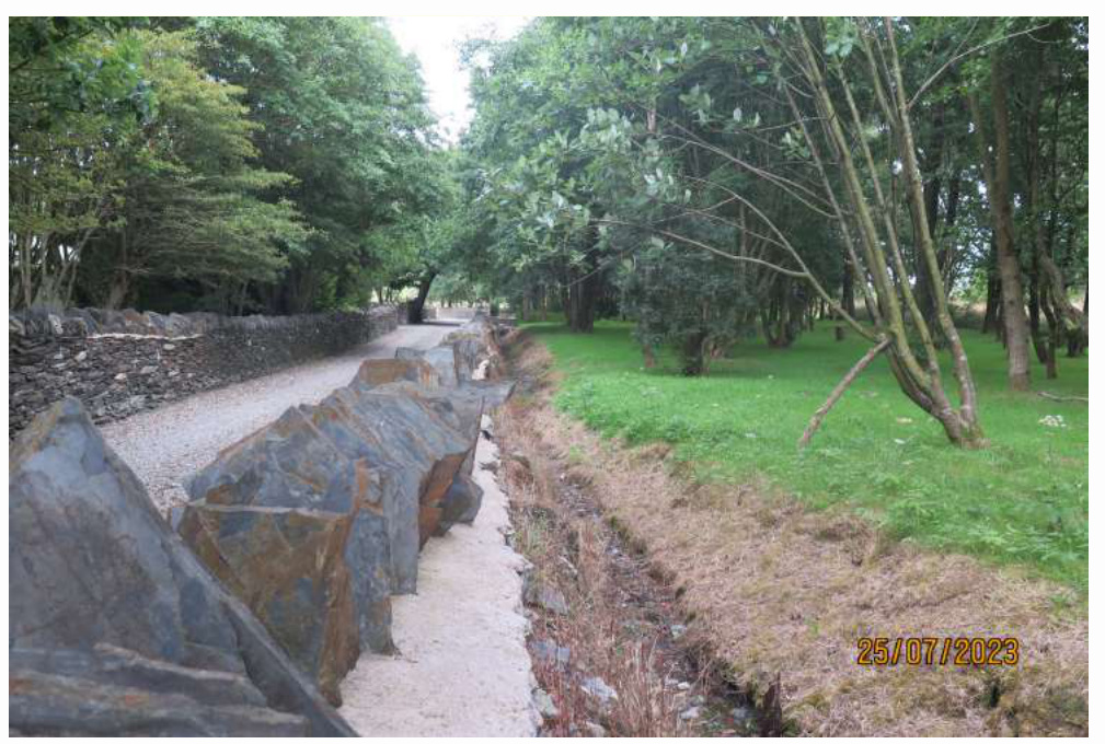 A photograph showing a gravel driveway bordered by a dry stone wall and large stone slabs in the foreground, with a grassy area and trees to the right.