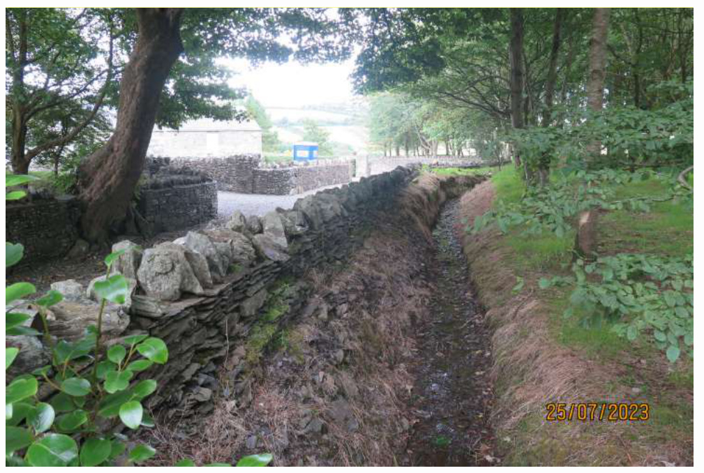 A photograph showing a long drainage trench dug alongside a traditional dry stone wall in a rural setting, with a white building visible in the background.