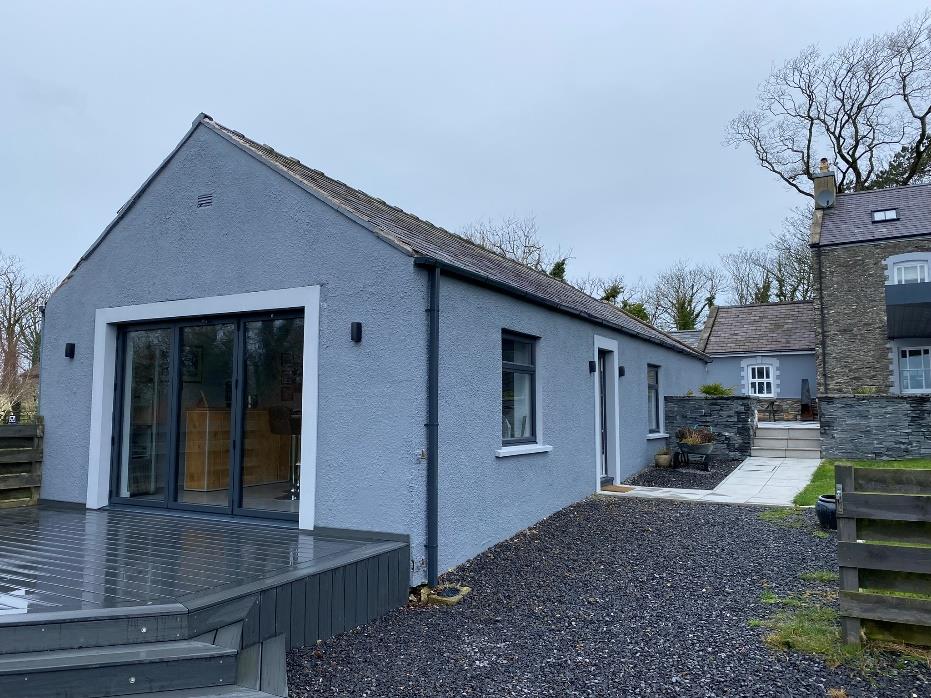 A photograph showing the exterior of a converted grey agricultural building with a modern wooden deck and large glass doors, situated next to a gravel driveway.