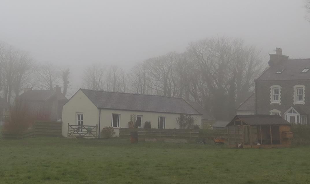 A foggy rural scene showing a long, single-story white building, likely a converted agricultural structure, situated next to a larger stone house.
