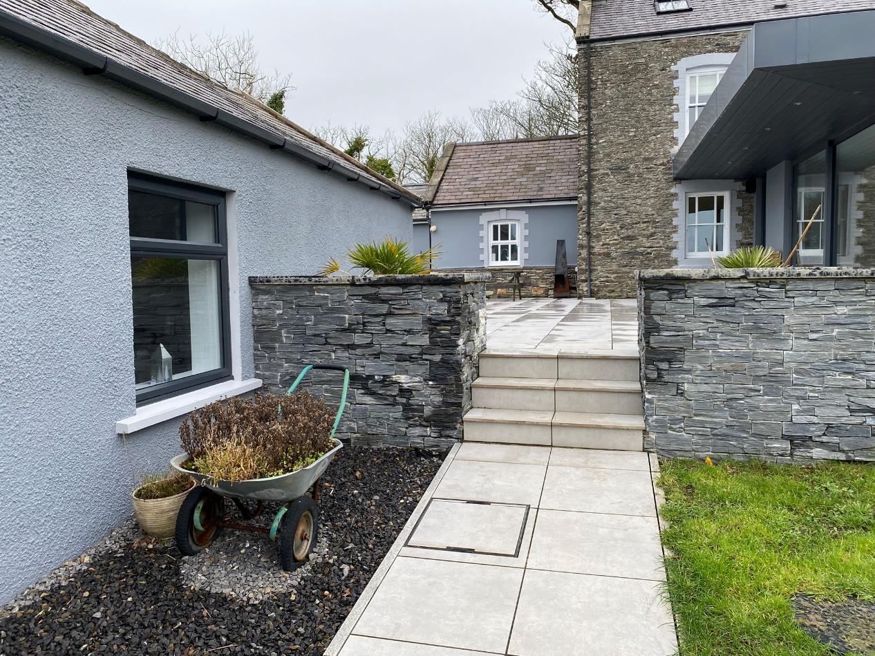A photograph showing the exterior of a property featuring a grey rendered building section on the left and a stone house on the right, connected by a paved patio with low stone walls.