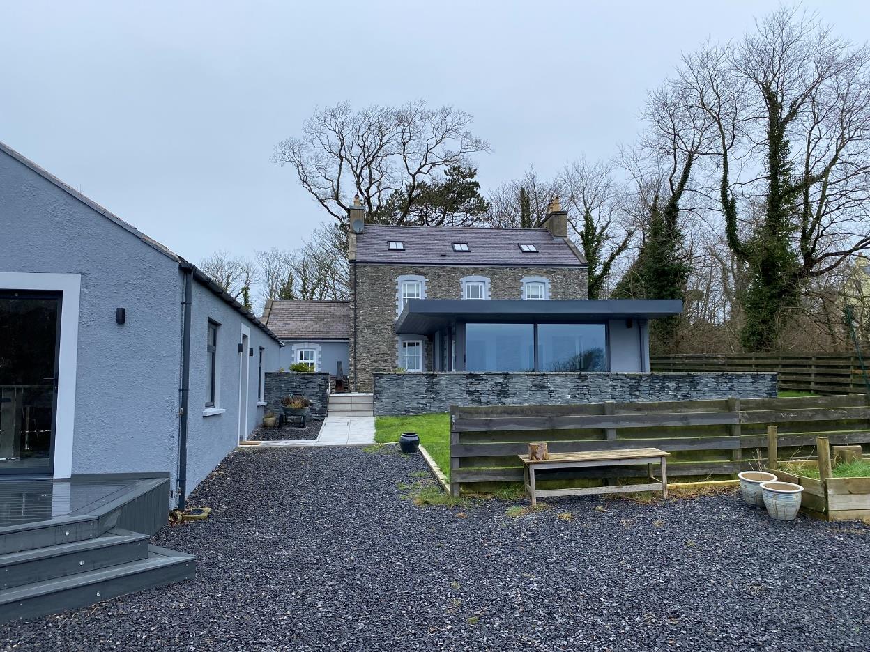 A photograph of a rural property featuring a traditional stone house and a modern grey single-storey extension with large glass windows.