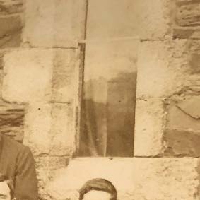 A sepia-toned historical photograph showing a stone wall with a window opening and partial views of people in the foreground.