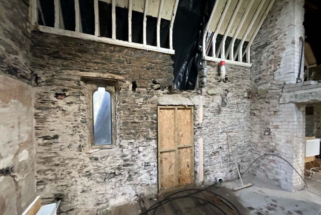 Interior photograph of a stone building undergoing renovation, showing exposed masonry walls, a boarded-up door, and a window opening.