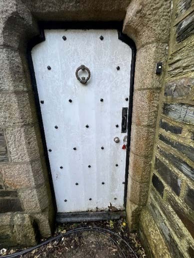 A close-up photograph of a white, studded wooden door with a ring handle set within a rough stone archway.