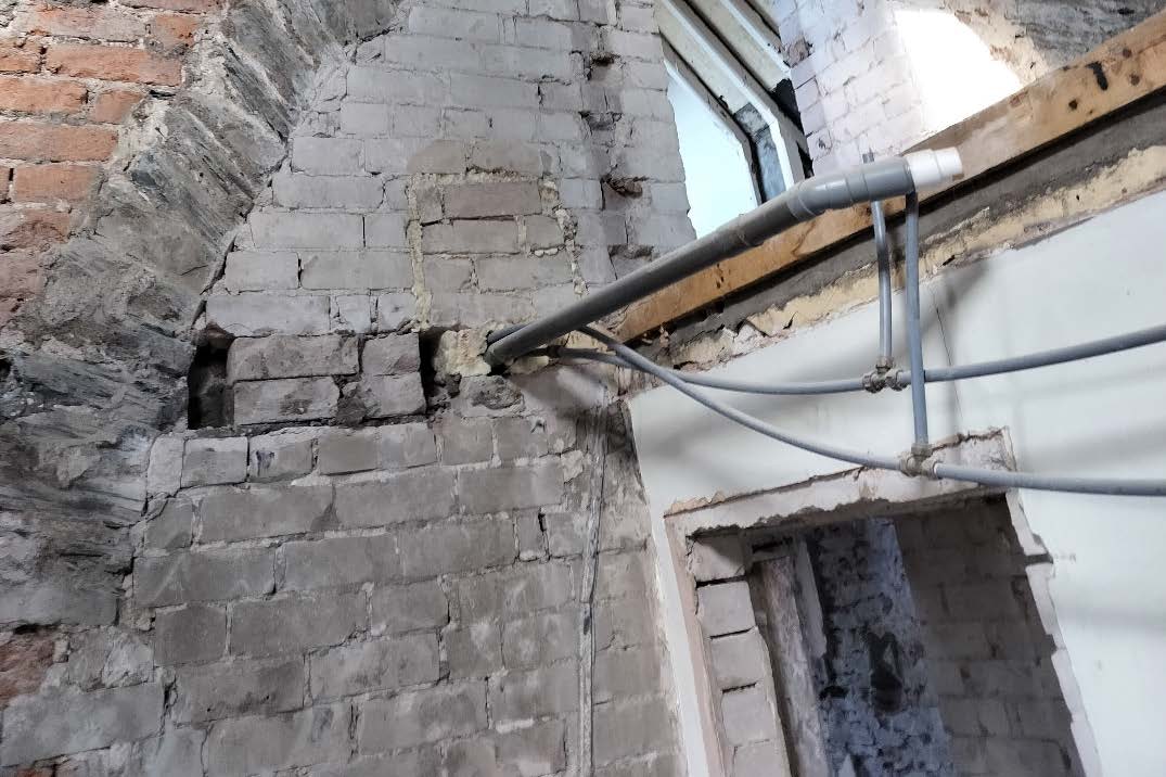 An interior photograph showing exposed grey brickwork, an arched structure, and grey drainage pipes running along a wall during renovation.